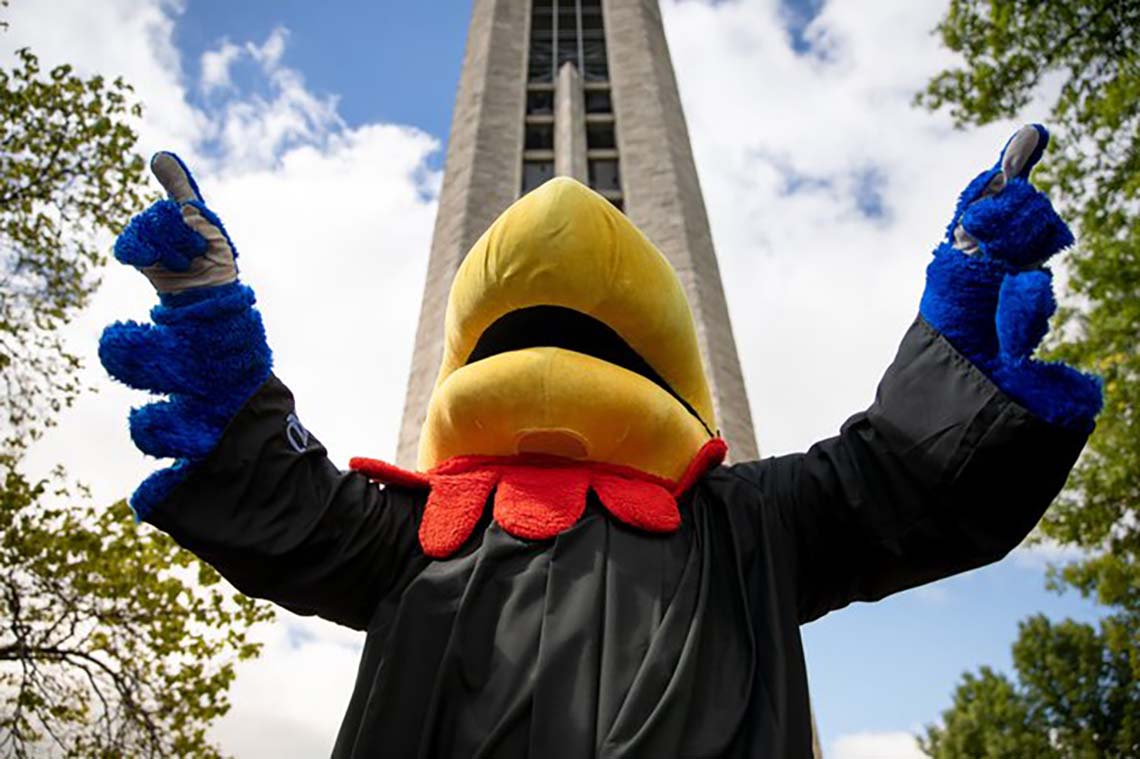 In front of the campanile, wearing a graduation gown, Big Jay stretches their wings in celebration