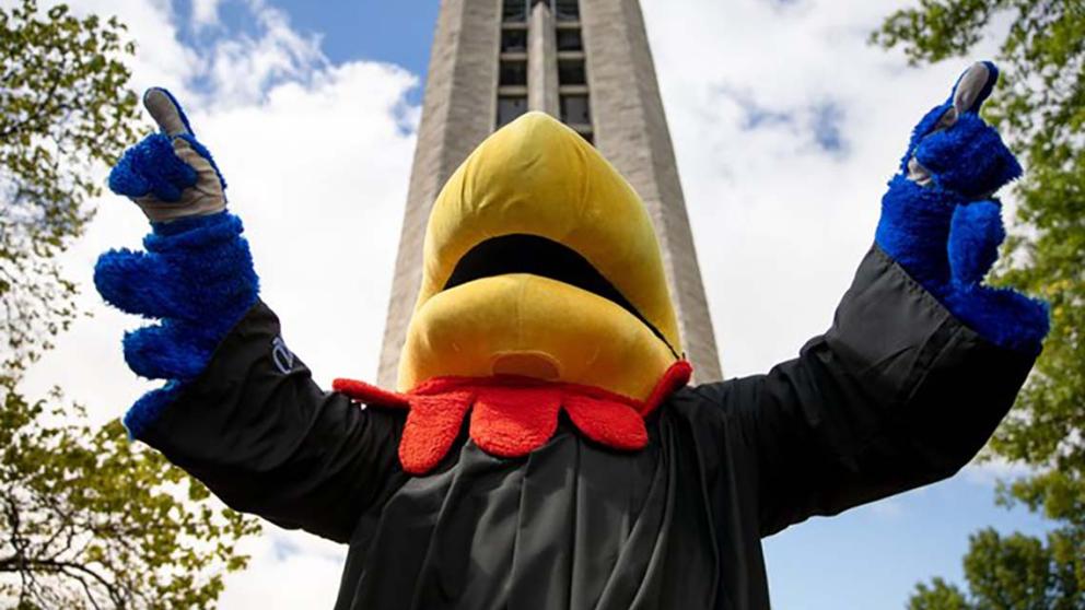 Big Jay, clad in a graduation gown, raises their arms in celebration in front of the campanile.